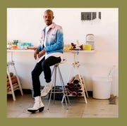 Diedrick Brackens - ELLE DECOR portrait of the artist sitting on a high stool at a workdesk and on the right a green sample of tapestry with a silouette at center