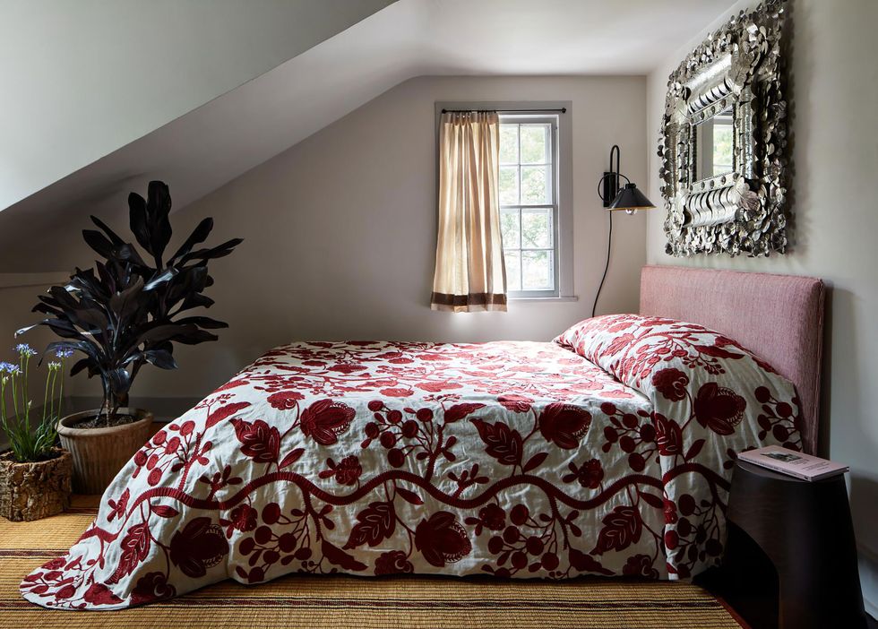 large bed with reddish pink upholstered headboard and a white coverlet patterned in bold red floral pattern in a bedroom with a sloped ceiling and a few plants in the corner and a silver framed mirror over the headboard on the wall