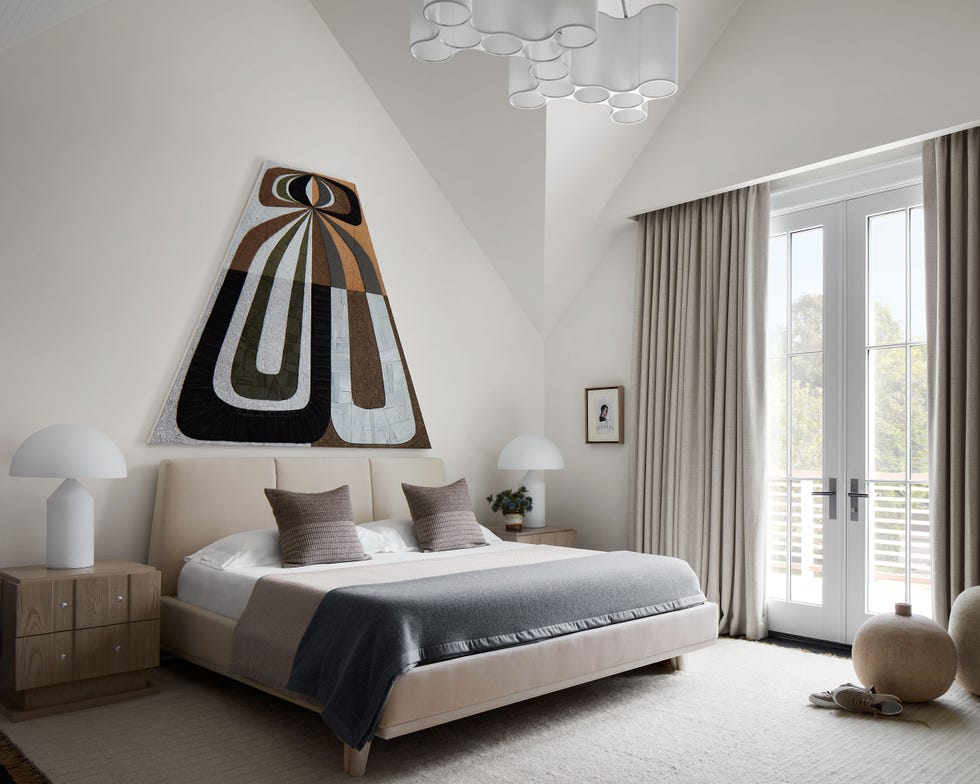 large bed with upholstered headboard and platform in ecru and gray with a piece of artwork in the shape of a trapezoid above it in the same tones and a light beige carpet and white mushroom lamps  on the bed stands on either side