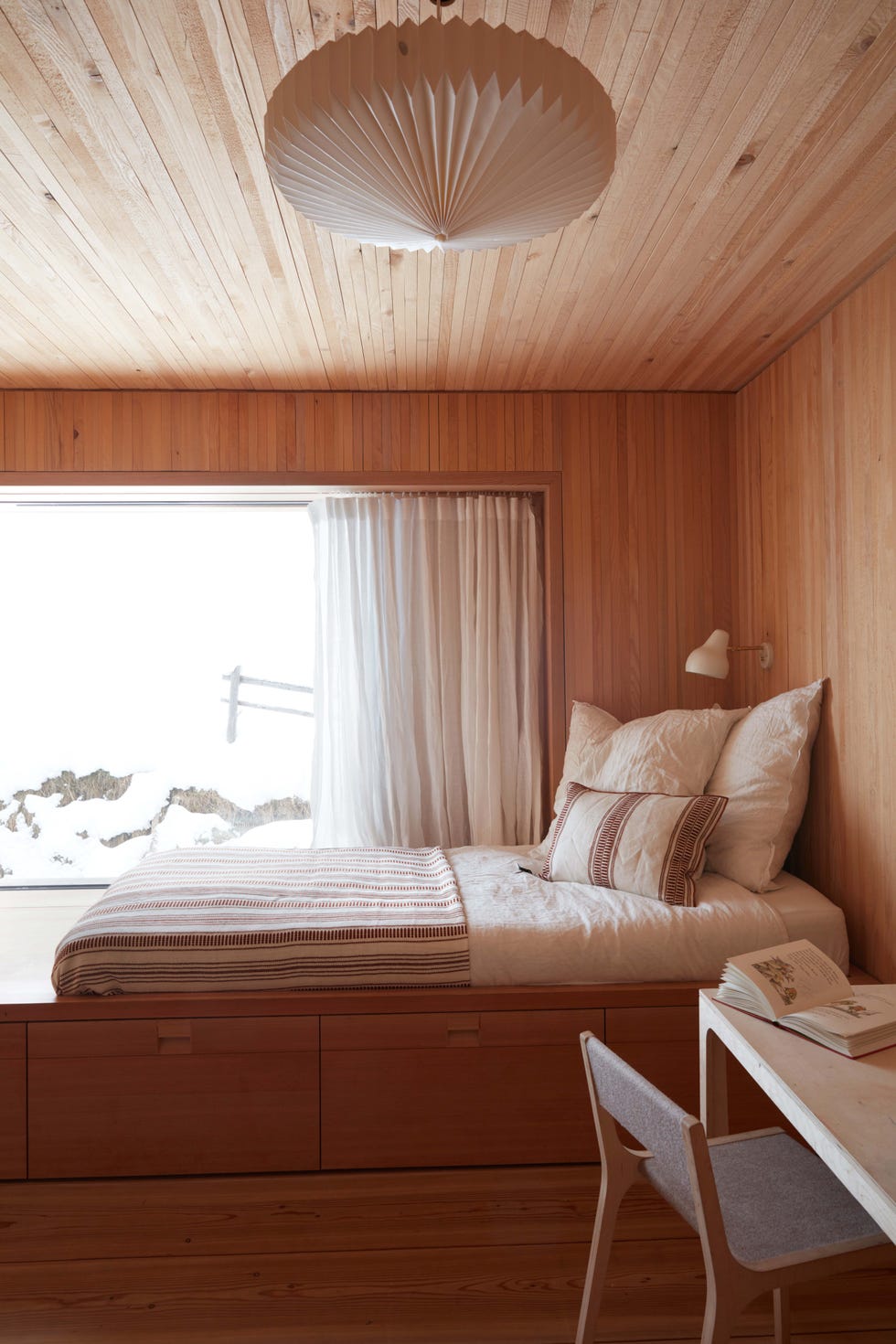 Guest Bedrooms - ELLE DECOR high platform bed with storage underneath and no headboard and room all in wood with striped linens and pillows on the bed and a small lamp overhead and a table and chair on one side and a large horizontal window with sheers opposite