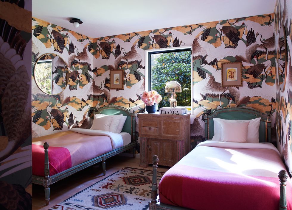 twin beds with bright pink and light pink linens and short acorn topped bedposts in a room with a colorful navaho type rug and very bold wallpaper in green and yellows that looks like geese wings and a wooden nightstand between the beds with a small window right behind it