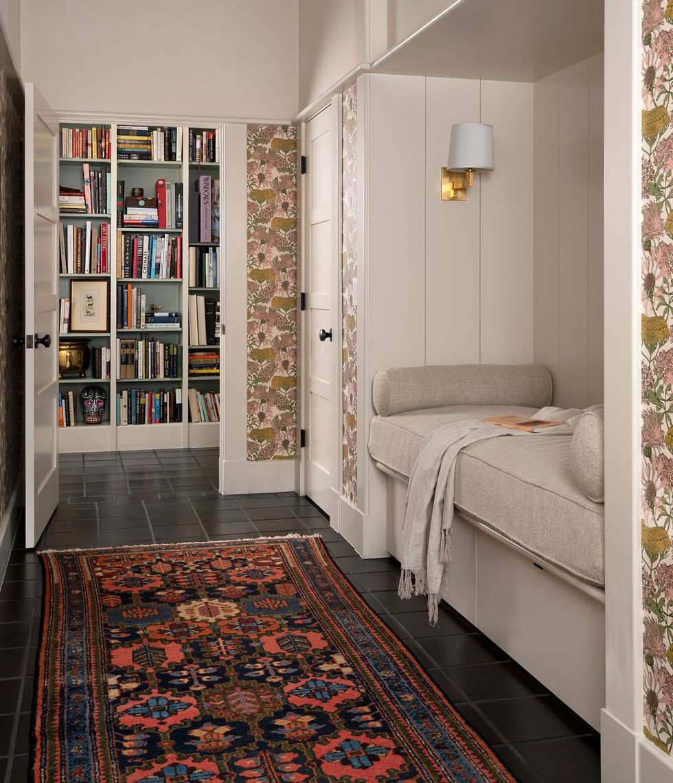 built in bed in ecru linen with raised platform and matching bolsters at foot and head and a flowered wallpaper in pink and ecru with a colorful carpet on dark floors and a floor to ceiling bookshelf seen through an open door into the next room