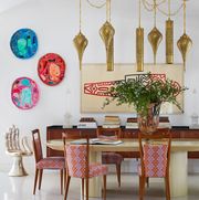 dining area with a long table with pinkish upholstered wooden chairs pulled up to it and multiple bronze fluted pendants hanging above it  on a chain and a silver hand chair in the background on the left next to a long console with stacks books atop and a large piece of artwork above it