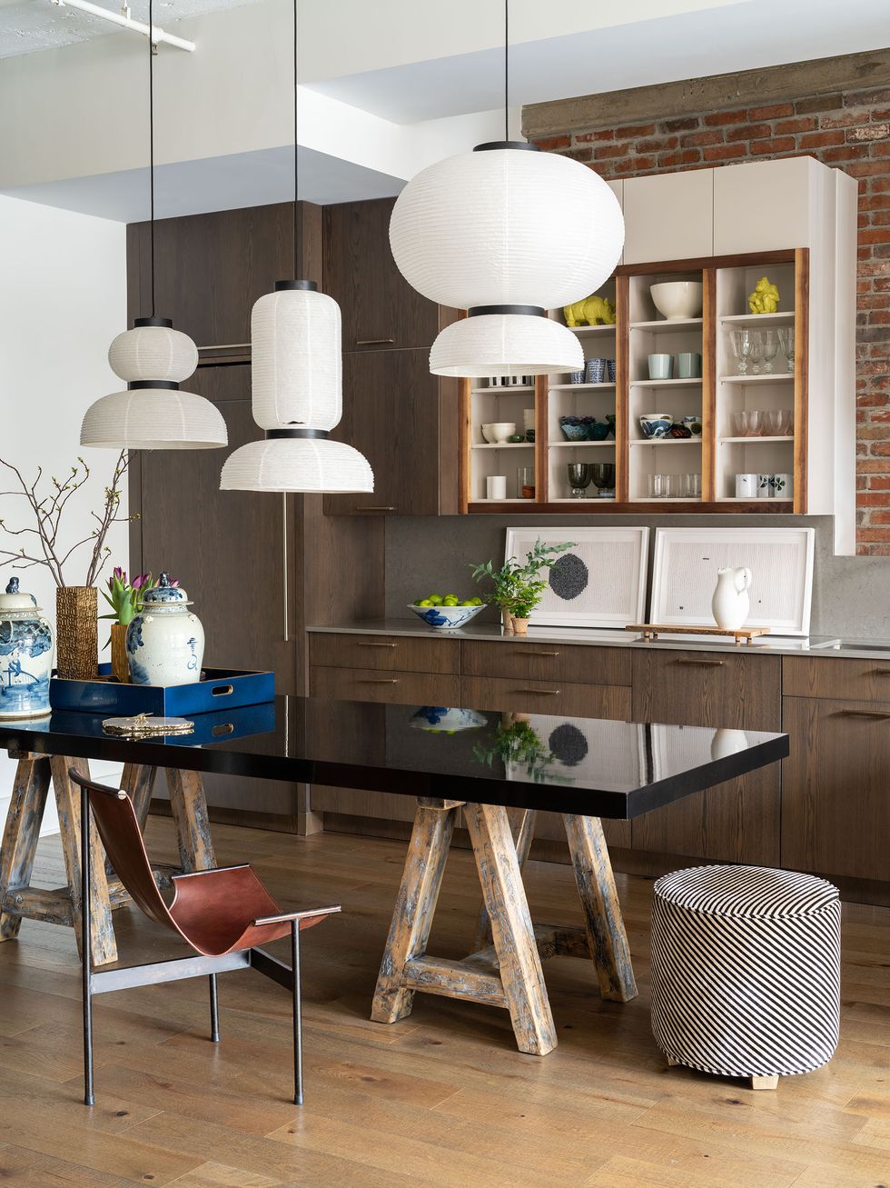 kitchen with a long shiny black table on distressed saw horse legs at center and a striped pouf and thin legged brown chair pulled up to it sleek modern counters are against the wall with framed prints on them leaning against the wall