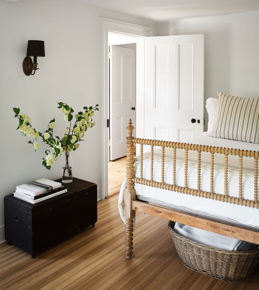 small bedroom with wooden floors and a wooden bed with a turned frame and white linens and a light green and yellow striped pillow and a basket underneath the bed and a dark low console opposite the bed next to the wall with a small vase with yellowish green flowers