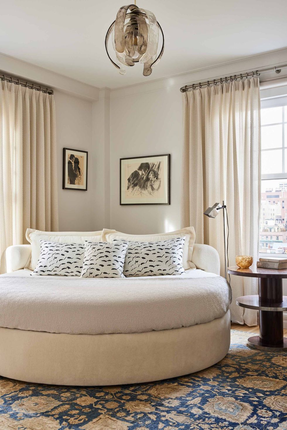 a round bed in a corner with black and white pillows and a two tier round nightstand and cream curtains on the tall windows and some framed artwork in the corner behind it and high pendant lamp