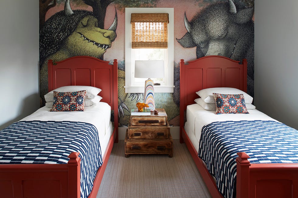 Guest Bedrooms - ELLE DECOR twin beds with dark red frame and high headboard with insets and blue and white pattern covers and a small stacked nightstand in between and a wallpaper on the wall behind the beds with figures from the where the wild things are