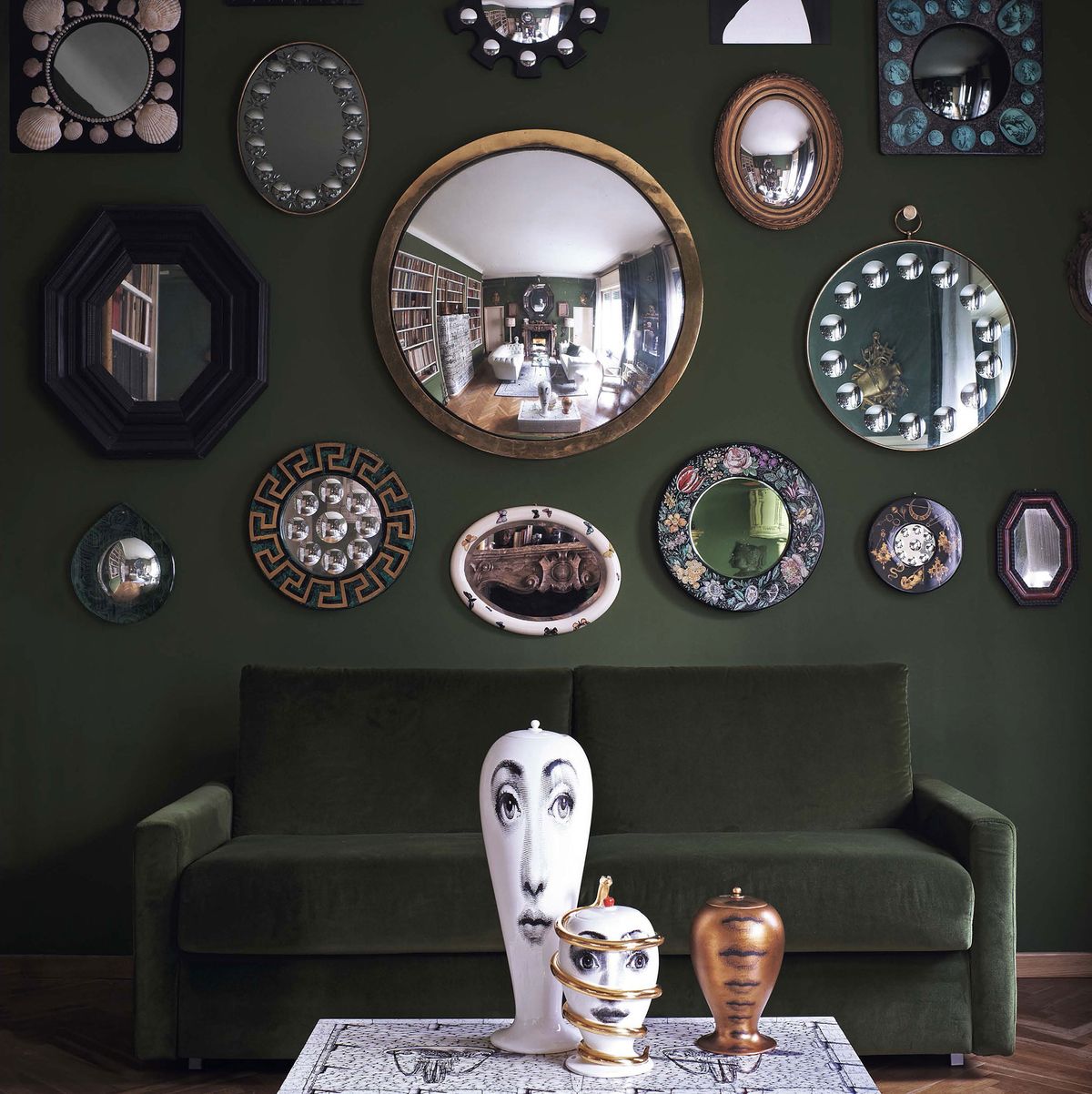 green velvet sofa against a dark green wall with round frames and mirrors of all sizes and shapes and on the low white cocktail table are classic fornasetti objects with faces on them