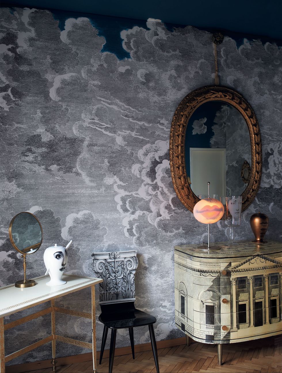 bedroom with dark wallpaper with a cloudy white print and a oval gilt mirror over a toile looking console and next to it a thing chair and table with classic objects with face designs on them