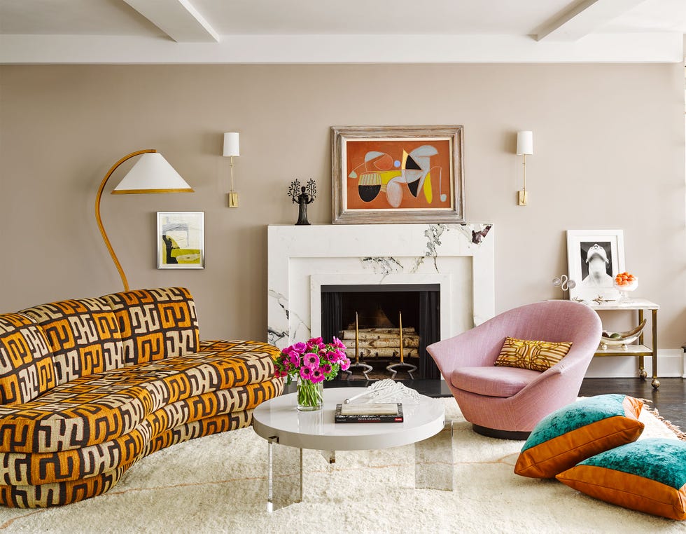 white marble mantel at center with rounded orange chaise to the left, white carpet, low round white table with clear legs, a pink mondern winged chair and pillows on the floor