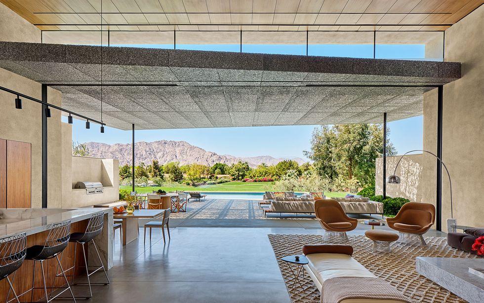 great room with counter and stools on left and sitting area with chairs and floor reading lamp on right opens onto large patio with chaise longues and in ground pool with trees and mountains beyond