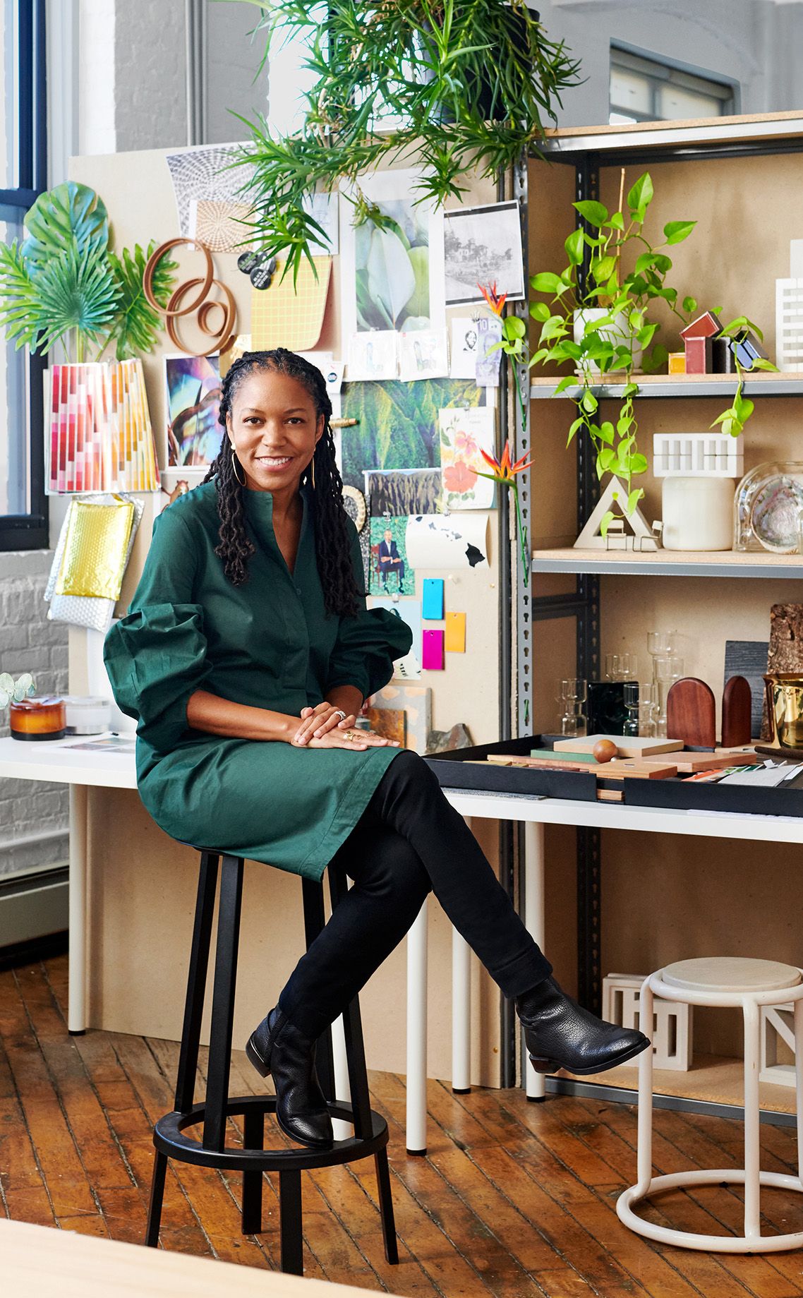 woman in green overdress and black pants sitting on a stool in front of a desk with patterns and samples and plants
