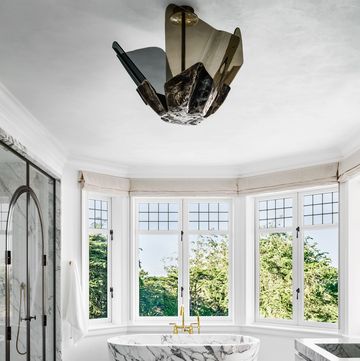 rounded bathroom with double windows in an arc at the back with a standalone marble tub at center and a floating marble vanity and a large bat like light fixture at center and a glass shower at left