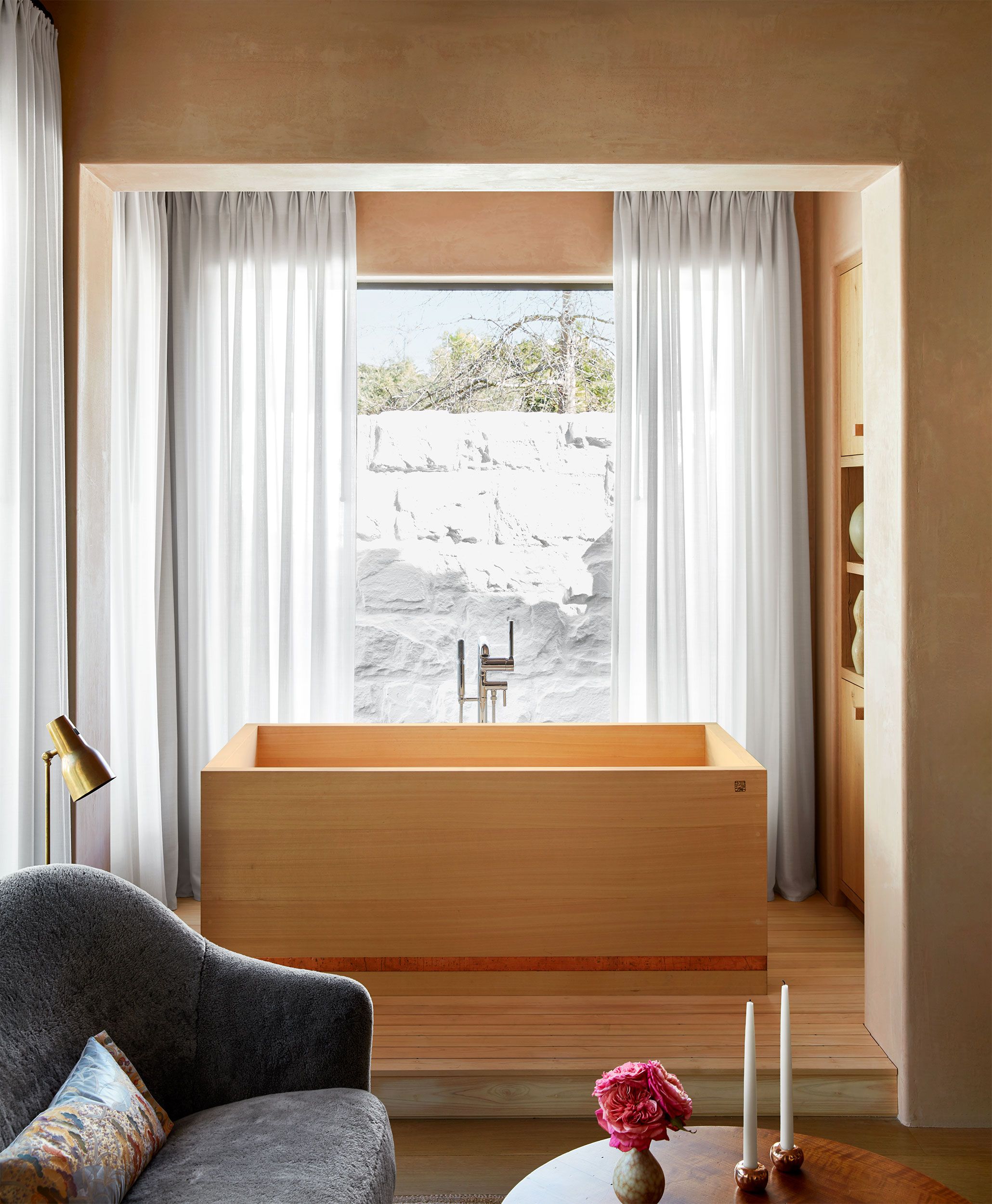 bathroom that looks like a room with a raised wooden platform on which is a wooden clad tub in front of the window and in the foreground is seen a sofa and small table with candlesticks