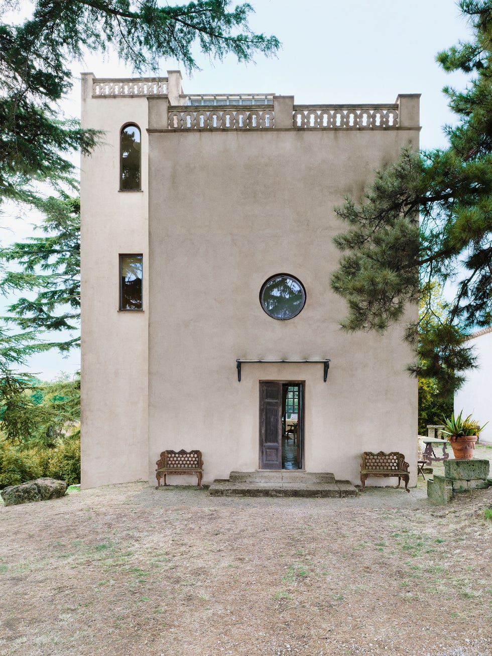 a tower with a stone colored plaster finish has stone steps and a wooden door with a circular window above it, two windows along its left side and a decorative rim around the rooftop, two cast iron benches flank the door