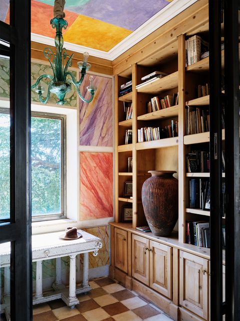 a library has shelving with books and a large vase, a low sideboard below a window surrounded by artworks in differently colored marble patterns, ceiling with brightly colored squares, and a green murano glass chandelier