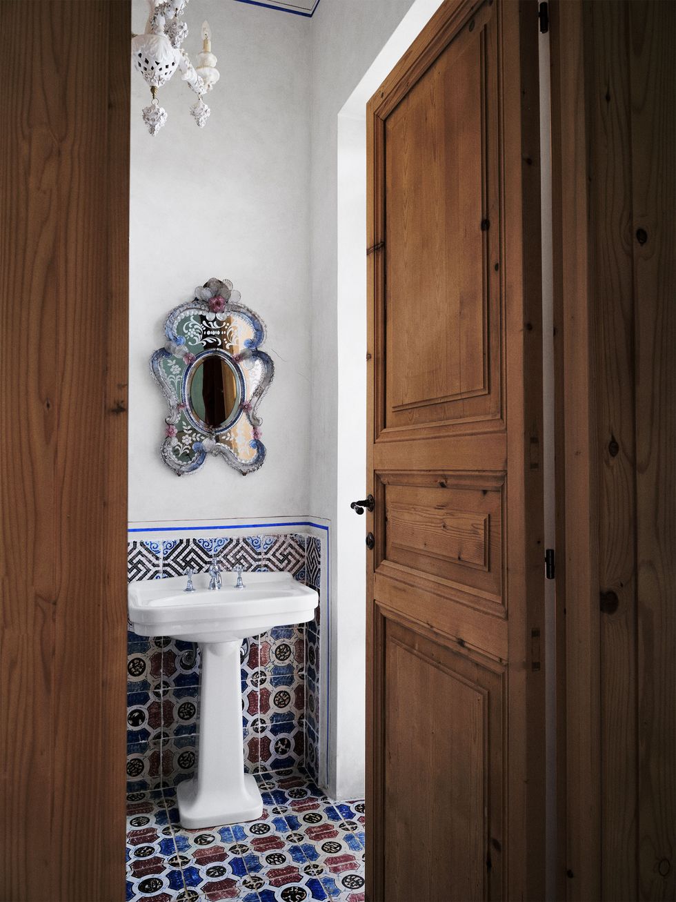a small bathroom with a carved wooden door has an 18th century murano mirror over a white pedestal sink with silver fittings and blue, black, red and white 19th century riggiole tile, and a white ceramic chandelier