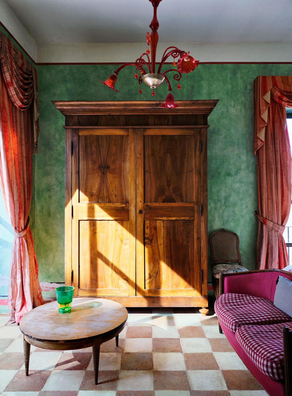 a guest bedroom has mottled green walls, a large wooden armoire, red silk curtained windows, a magenta sofa with checked seats, an oval wooden cocktail table, a red murano glass chandelier, and checkered tile floor