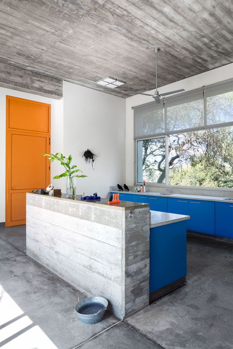 La cuisine a des sols en ciment gris, deux ensembles d'armoires bleu foncé surmontées de comptoirs en ciment poli, un juste en dessous des fenêtres, une porte orange vif à une extrémité et un ventilateur de plafond.