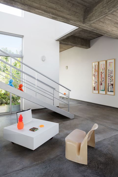 a living room's walls and a staircase are a light gray, the floor is a darker cement, three vertical framed artworks are on the wall facing the stairs, a low square cocktail table and molded plastic chair sit near stairs