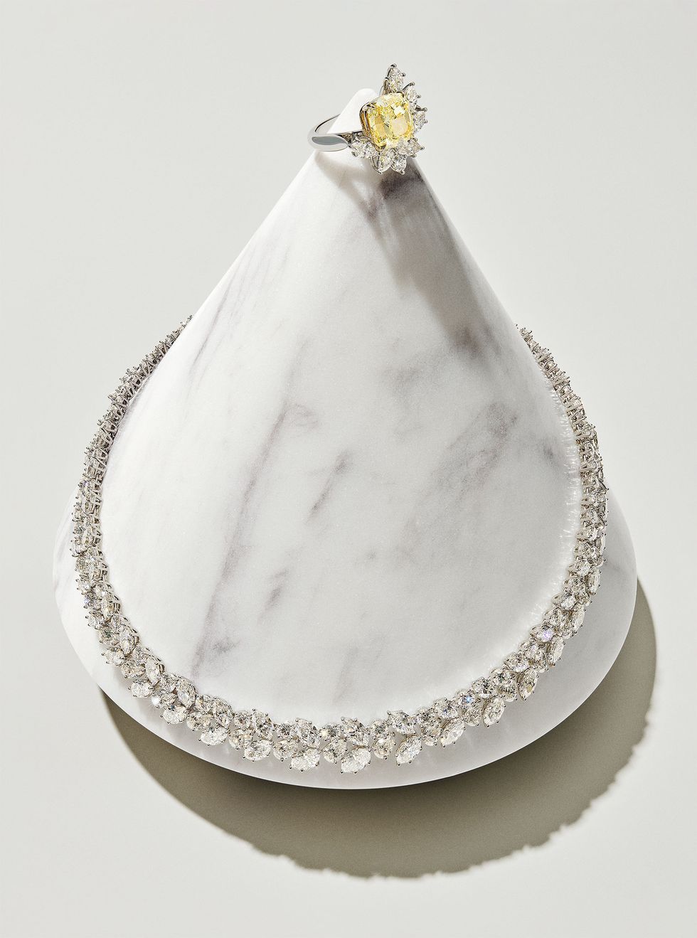 cushion cut yellow diamond ring and diamond cluster necklace displayed on a marble cone with the ring at the pointed top