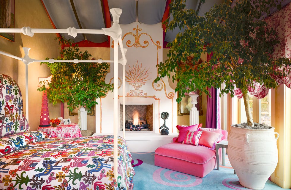 metal four poster bed painted white, bright print bedcover, pink lounge chair, blue rug with pink swirls, fireplace with floor to ceiling decorative surround, tented ceiling, urn with small tree, yellowish walls