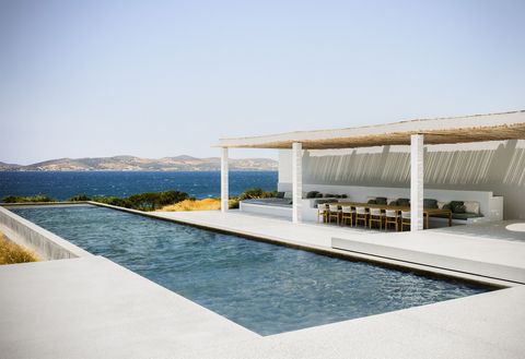 a concrete terrace surrounds a rectangular pool on three sides, to the right is a pergola of wood beams above a dining table with seven chairs and built in cushioned benches facing them, a lounge area is at the end of table