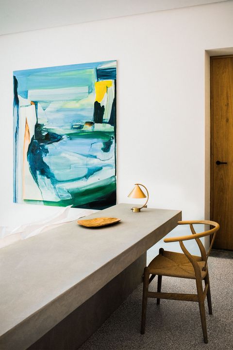 a guest bedroom has white pillows propped against a grayish microcement headboard that doubles as a writing desk, which has a chair and a small lamp, a primarily blue and green abstract painting is on the wall