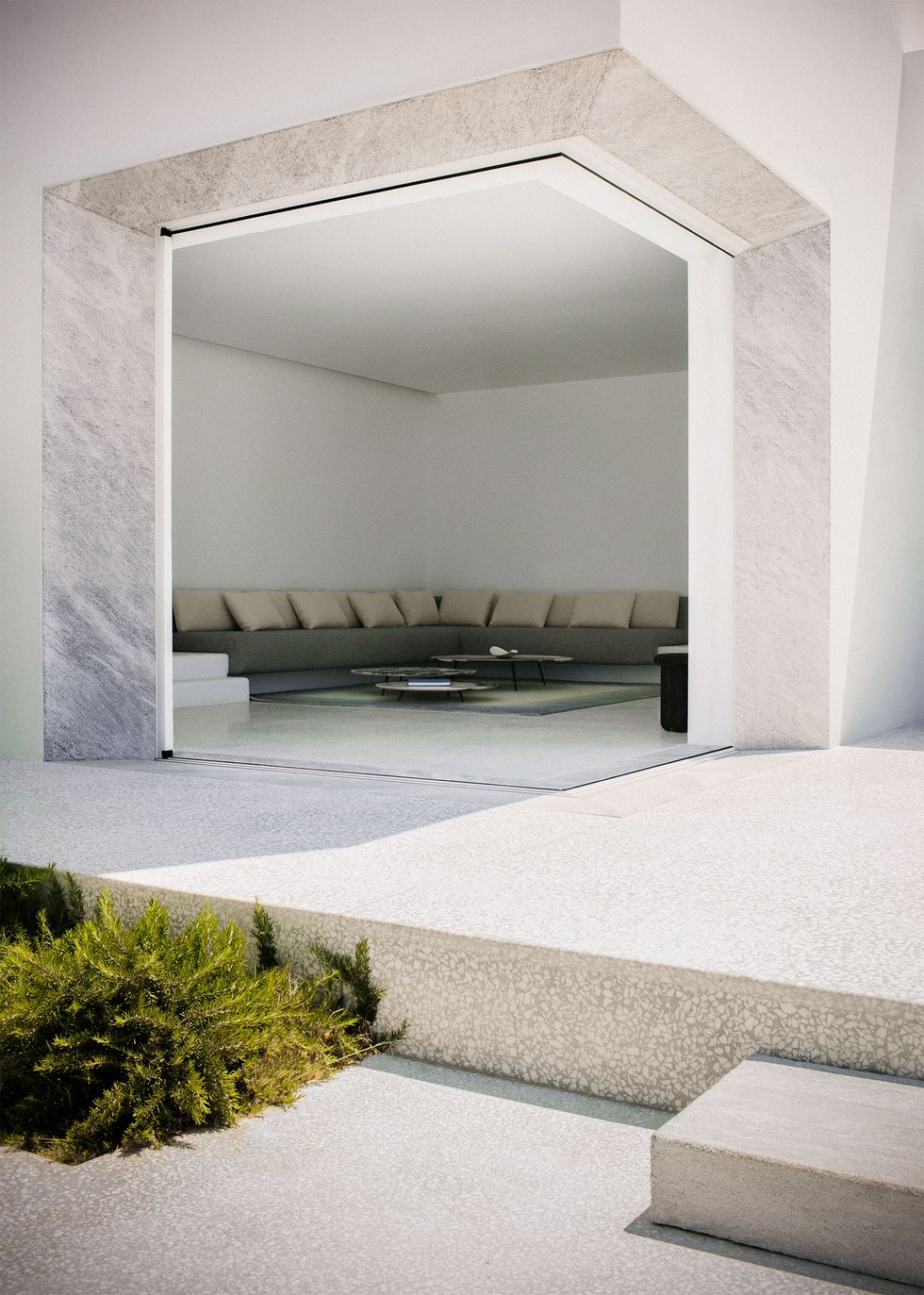 from the exterior, a single step up onto the terrace leads to an indoor living room with a large built in l shaped sofa with cushioned seats and backs and pillows and three low cocktail tables of varying sizes