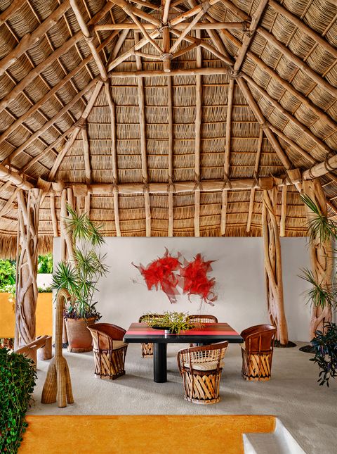 in a thatched dining palapa there is a square black table with red and other colored stripes on top, five barrel chairs, a standing woven sculpture, a red wire mesh wall artwork and some plants in pots