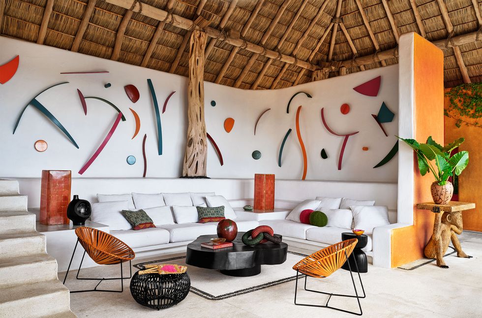 in a white living room are built in side tables and seating with colored accent pillows, a dark wood curved cocktail table, woven seat side chairs, and multiple colored shapes made of painted stainless steel on the walls