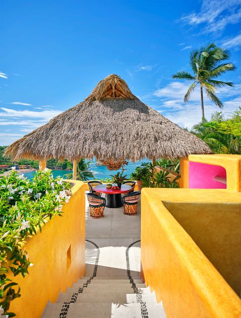 open air steps embedded with stones and yellow walls lead to a dining palapa with a resin topped round table, four leather backed barrel chairs, and a toucan parrot monkey chandelier, with the cove in the background