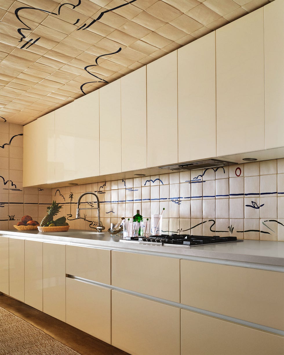 Martin Brule - ELLE DECOR a kitchen with glossy ivory colored cabinets above and below a light counter with a stovetop and sink backsplash wall and c
