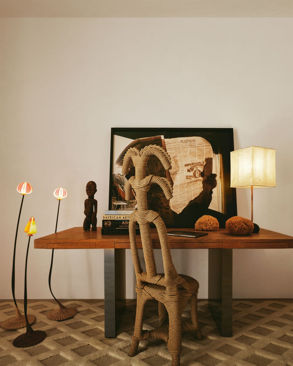 home office with a walnut desk top and steel legs with a small sculpture of a person, lamp with paper shade, small coral pieces, books and an artwork, vintage rope chair, three small shell floor lamps, textured rug