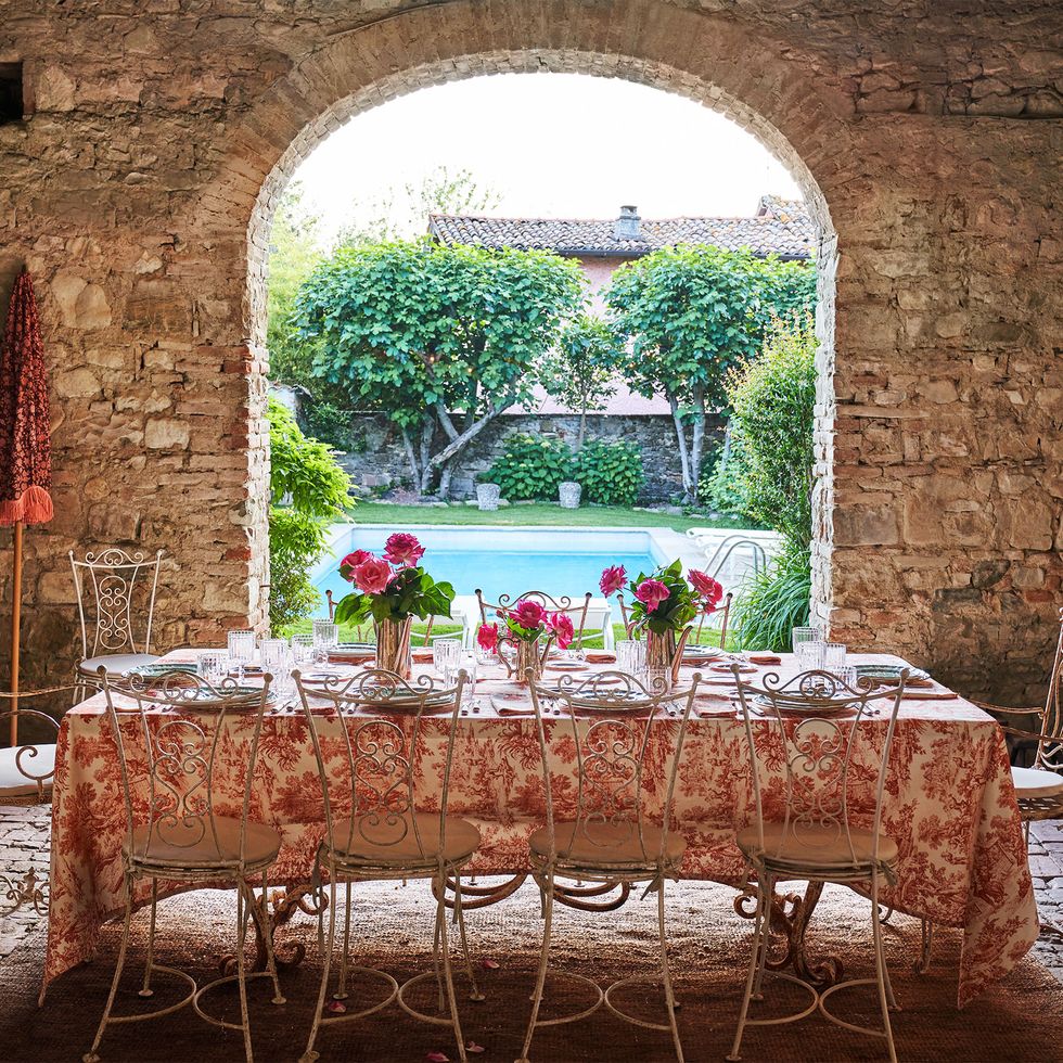 a poolside room has a terra cotta floor and rustic stone walls with an arched opening to the pool, inside is a cloth covered dining table with place settings and cast iron chairs with open ornamental backs