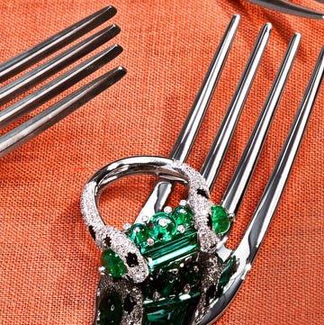 a table covered in woven terra cotta colored cloth with four different shaped silver forks, one of them with a ring of platinum, ﻿emeralds, onyx, ﻿and diamonds
