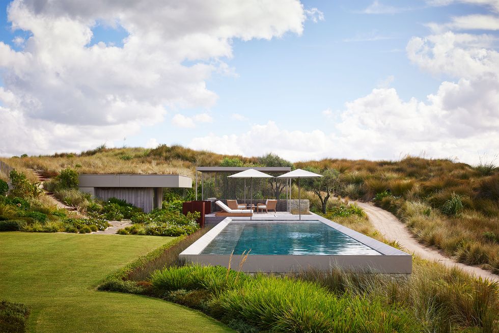 Next to a dirt road is a swimming pool with umbrellas and chairs next to a pool house built into the grassy hillside