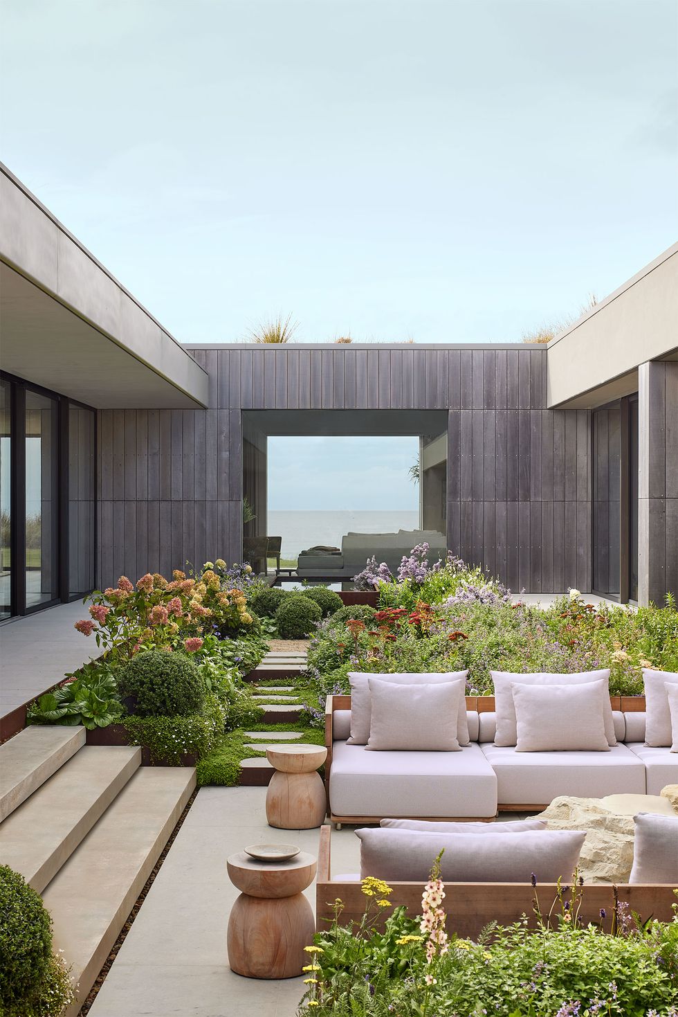 A patio framed in gray wood with opposing wooden sofas with white cushions, two circular wooden tables, steps leading inside and to a garden area with shrubs and flowers, and a large window overlooking the sea.