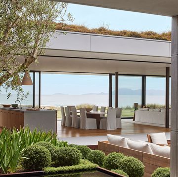 through a courtyard that has a tree and shrubs and a sofa is a view of a large open room with a kitchen island, a dining table with white cloth covered chairs and a sofa and glass wall with a sea and mountain views beyond