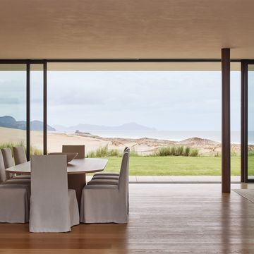an oval dining table with 10 chairs covered in white fabric, wood flor, glass walls looking out to the grass lawn, sand dune, and the sea and mountains beyond