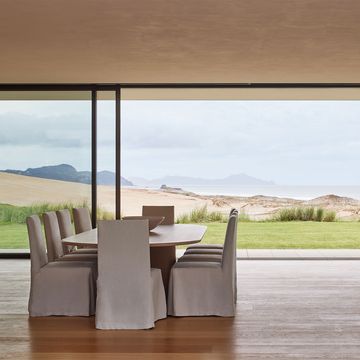 an oval dining table with 10 chairs covered in white fabric, wood flor, glass walls looking out to the grass lawn, sand dune, and the sea and mountains beyond