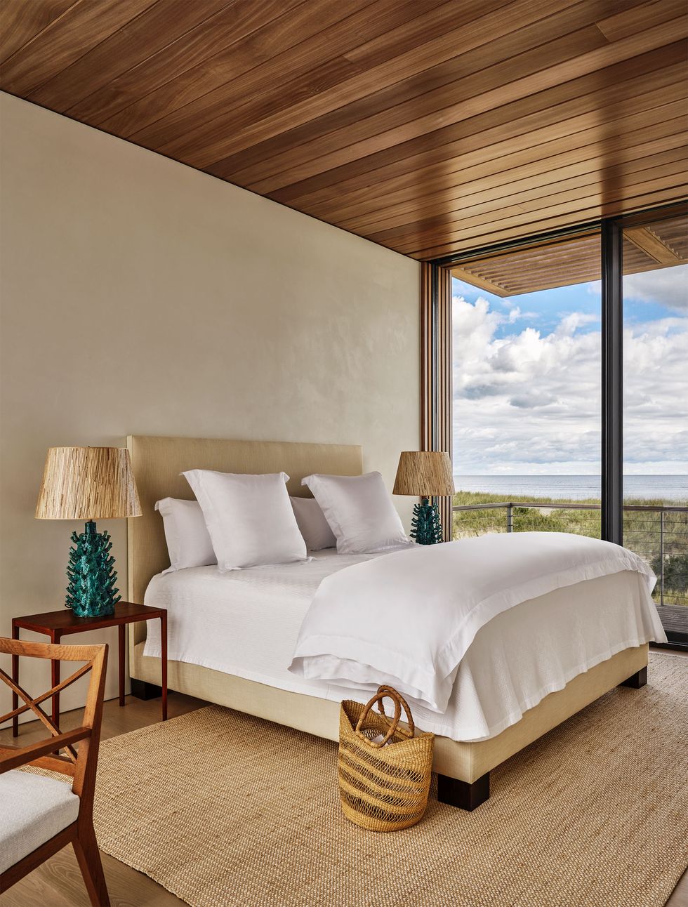 Bed with upholstered headboard and frame, white bedding, night tables with lamps with coral-like bases, natural rug, bedside basket, wooden ceiling and floor, glass doors to the terrace above the sand dunes