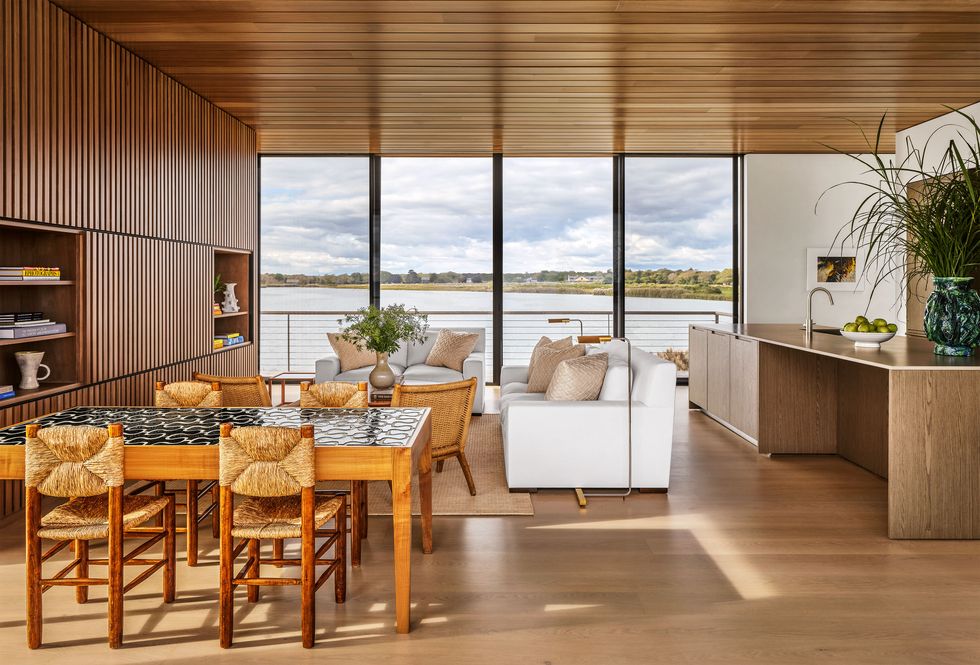 Large open room with glass wall overlooking the sea, dining table and chairs, sofa, double chair, cocktail table, wooden slatted wall with built-in shelves, kitchen island with sink and cabinets, wooden ceiling and floor
