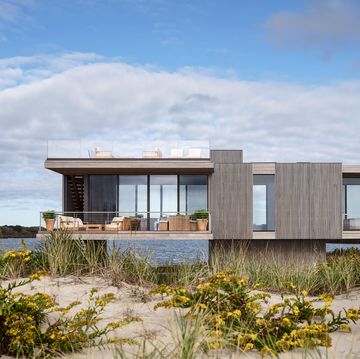 a rectangular house with wood panels and glass walls perches on a base in grassy dunes next to the sea, lounge furniture on a terrace on first floor, rooftop terrace lounge