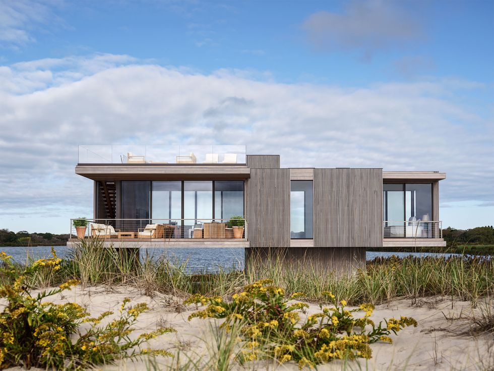 a rectangular house with wood panels and glass walls perches on a base in grassy dunes next to the sea, lounge furniture on a terrace on first floor, rooftop terrace lounge