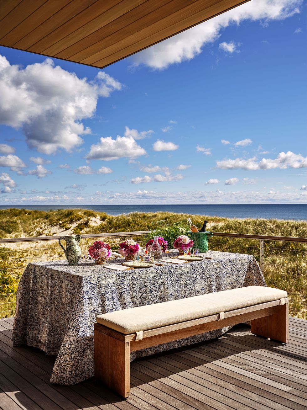 Wooden deck and wire railing overlooking the grassy dunes and sea, table with plates and flowers, printed tablecloth, bench with seat cushion