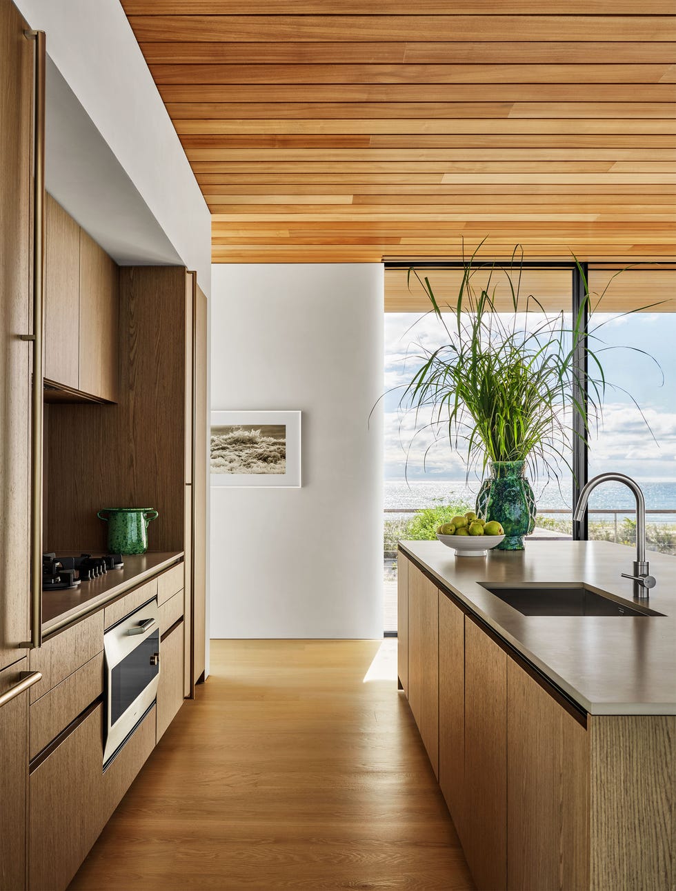 Kitchen with built-in stove and oven on wood counters and cabinets, kitchen island with sink and cabinets, wood floors, white walls with framed artwork, and glass walls with ocean views.