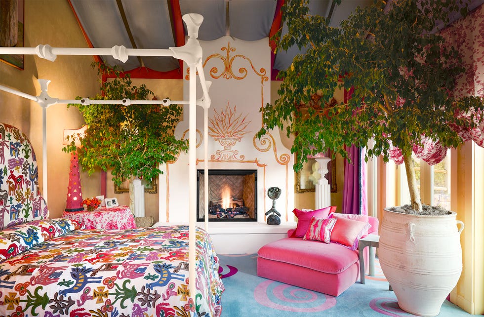 metal four poster bed painted white, bright print bedcover, pink lounge chair, blue rug with pink swirls, fireplace with floor to ceiling decorative surround, tented ceiling, urn with small tree, yellowish walls