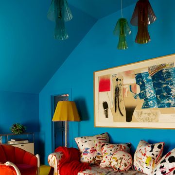 a study with bright blue walls, multicolored sofa with pillows, two red upholstered chairs with swan necks as arms, painted hexagonal table, floor lamp, large artwork, three glass pendants, mustard rug