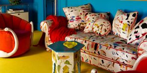 a study with bright blue walls, multicolored sofa with pillows, two red upholstered chairs with swan necks as arms, painted hexagonal table, floor lamp, large artwork, three glass pendants, mustard rug